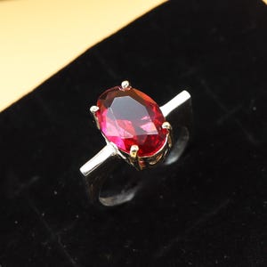 May include: A silver ring featuring a large, oval-cut, deep red gemstone. The gemstone is set with four prongs, and the band is a simple, polished silver. The ring is displayed against a black background.