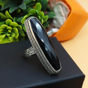 May include: A silver ring with a large, elongated black gemstone. The gemstone has a faceted cut, and the ring features an ornate, detailed design around the stone and on the band. The ring is set against a black surface.
