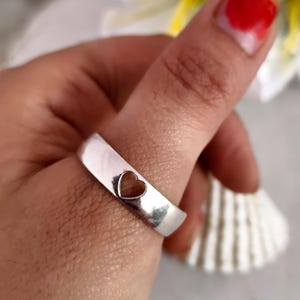 Peut inclure: Une bague en argent avec un cœur évidé. La bague a une surface lisse et brillante. Le cœur est centré sur le groupe. La bague est portée sur un doigt.