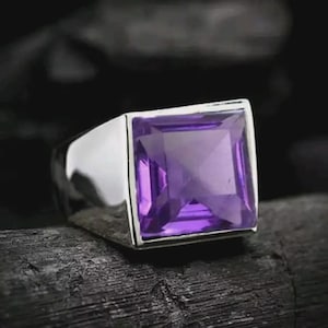 May include: A silver ring with a large, square-cut, purple amethyst gemstone. The ring has a polished, shiny finish and a thick band. The gemstone is set in a square bezel, and the ring is displayed against a dark background.