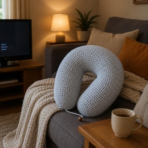 May include: A light blue, U-shaped crochet travel pillow rests on a gray sofa. The pillow has a textured surface and a wooden bead detail. A beige blanket and orange pillow are also on the sofa. A cup of tea sits on a wooden table.