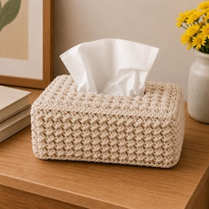 May include: A rectangular tissue box cover in a light beige crochet pattern. White tissues are pulled from the top opening. The box is on a wooden surface, with a vase of yellow flowers in the background.