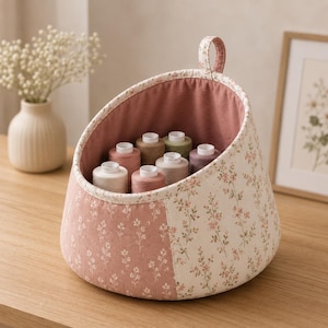 May include: A fabric storage basket with a slanted opening and a top hanging loop. The exterior features a two-tone floral pattern in dusty pink and cream, while the interior is lined with solid dusty pink fabric. It holds seven spools of thread.