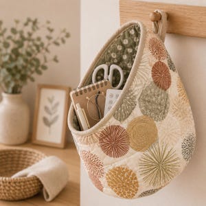 May include: A quilted fabric hanging storage basket featuring a pattern of circular dandelion-like motifs in shades of rust, mustard, and sage green. The basket contains a small spiral-bound notebook, a white pen, glasses, and white scissors.