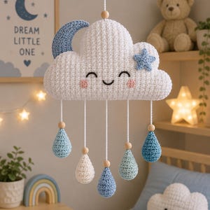 Op de afbeelding: Een handgemaakte gehaakte wolkenmobiel met een lachend gezicht, een blauwe halve maan en steraccenten. De wolk heeft hangende druppels in verschillende tinten blauw en wit. De kinderkamerdecoratie omvat een ingelijste print met de tekst "Dream Little One".