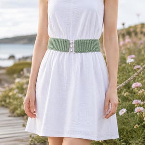 May include: A white sleeveless dress featuring a textured fabric and a sage green ribbed crochet belt. The belt is secured at the center with a silver-toned metal clasp that features three small, circular, white decorative accents in a vertical row.