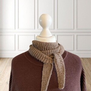 May include: A brown knitted scarf tied in a knot, displayed on a mannequin. The scarf is a light brown color and has a ribbed texture. The mannequin is wearing a brown sweater. The background is a white wall with paneling.
