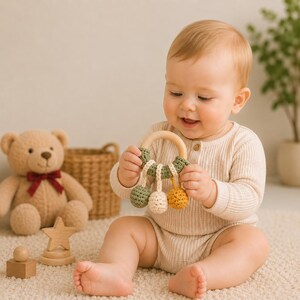 May include: A wooden ring baby rattle featuring a sage green crocheted cover and three dangling crocheted spheres in sage green, cream, and mustard yellow. The rattle is held by a baby wearing a beige ribbed long-sleeve romper.