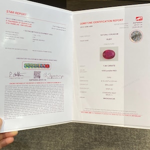 Könnte beinhalten: Ein Edelstein-Identifikationsbericht des GFCO Gemological Laboratory für einen ovalen, natürlichen Korund-Rubin mit 1,44 Karat. Das Dokument enthält Angaben zu lebhaft purpurroter Farbe, Brillantschliff und Herkunft aus Madagaskar.
