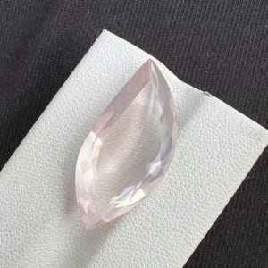 Peut inclure: Une pierre précieuse facettée et allongée, de couleur rose pâle. La gemme est posée sur une surface blanche et texturée, et la taille de la pierre crée de multiples surfaces réfléchissantes. La forme est similaire à une feuille ou une goutte d'eau.