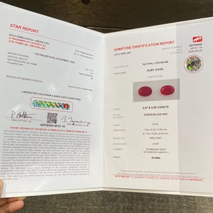 Könnte beinhalten: Ein GFCO gemmologischer Laborbericht für ein Paar natürlicher Korund-Rubine. Das Dokument beschreibt die taubenblutrote Farbe, die ovale Form, den Brillantschliff und die Gewichte von 0,97 und 0,89 Karat, mit Abmessungen in Millimetern.