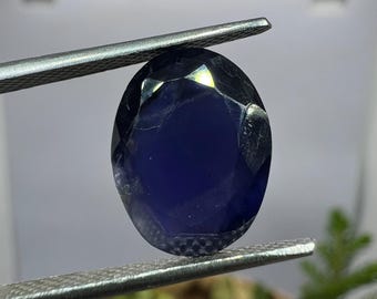 May include: A faceted, oval-shaped, deep blue gemstone is held by silver tweezers. The gem has a polished surface with multiple facets, reflecting light. The background is blurred, with hints of green foliage.