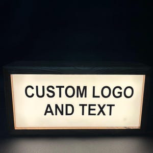 May include: A rectangular illuminated sign with the text "CUSTOM LOGO AND TEXT" in bold, black letters. The sign has a wooden frame and emits a soft, warm light against a dark background. The sign is designed for custom branding.