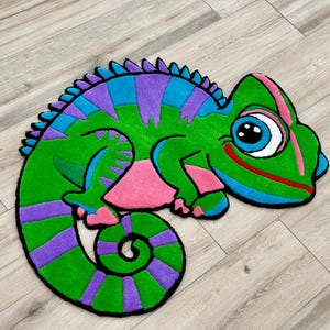 Puede incluir: Una colorida alfombra con forma de camaleón de dibujos animados. La alfombra presenta un cuerpo verde con rayas moradas y azules, una panza rosa y una cola enroscada. El camaleón tiene un ojo grande y una boca roja sonriente.