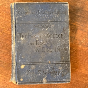 May include: A vintage blue book titled "Elementary Lessons in English" with the text "The Parts of Speech and How to Use Them." The book is worn, with visible signs of age and use. The publisher's name, Ginn & Company, is also visible.