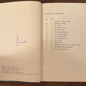 May include: An open book showing the table of contents for a biography of Theodore Roosevelt. The page lists chapter titles such as "Weak Boy, Strong Man" and "The Wild Life". The book is open on a wooden surface.