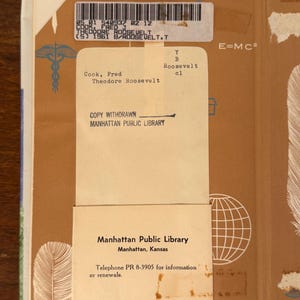 May include: An open book with a tan cover and a library card. The card states "Manhattan Public Library" and a phone number. The book's spine has a barcode and the title "Theodore Roosevelt". The cover has a feather and globe graphic.