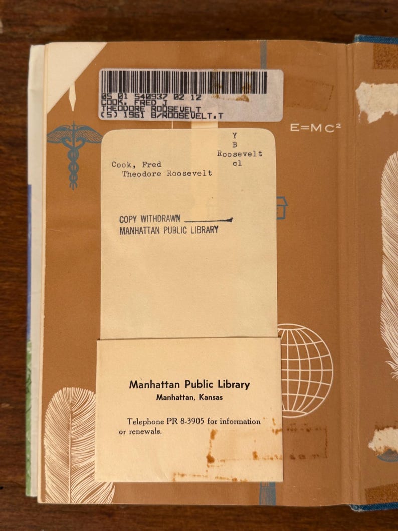 May include: An open book with a tan cover and a library card. The card states "Manhattan Public Library" and a phone number. The book's spine has a barcode and the title "Theodore Roosevelt". The cover has a feather and globe graphic.