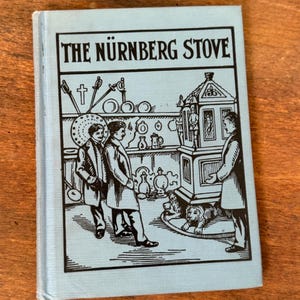 May include: A small, light blue book titled "THE NÜRNBERG STOVE" in black text. The cover features a black and white illustration of several figures and a stove. The book is on a wooden surface.