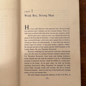 May include: An open book showing the start of a chapter titled "Weak Boy, Strong Man". The text is black on a cream-colored page. The background is a wooden surface. The text describes a biographical narrative.