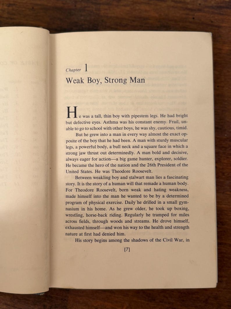 May include: An open book showing the start of a chapter titled "Weak Boy, Strong Man". The text is black on a cream-colored page. The background is a wooden surface. The text describes a biographical narrative.