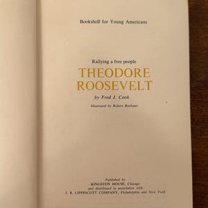 May include: An open book displaying the title page of "Theodore Roosevelt" by Fred J. Cook. The book is from the "Bookshelf for Young Americans" series. The title and author's name are in a gold font.