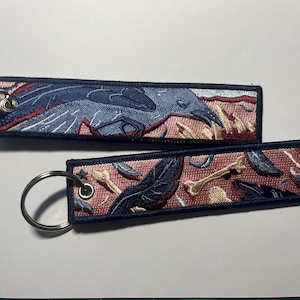 Puede incluir: Dos llaveros bordados con borde azul oscuro. Uno presenta un diseño de pájaro azul con detalles en rojo y blanco. El otro tiene un patrón de huesos y plumas en azul y beige sobre un fondo rosa.