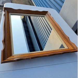 May include: A rectangular mirror with a decorative, light brown wooden frame. The frame has a scalloped edge design. The mirror reflects a building with vertical lines and a white surface.