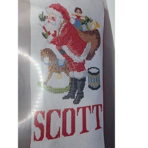 May include: A white fabric Christmas stocking featuring a cross-stitch design of Santa Claus carrying a brown sack filled with toys, a brown rocking horse, and a blue drum. The name SCOTT is embroidered in large red capital letters at the bottom.