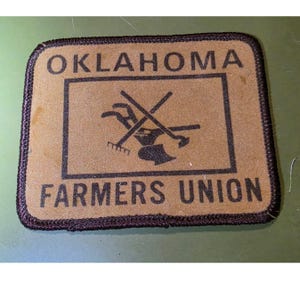 May include: A rectangular patch with the words "OKLAHOMA FARMERS UNION" in black block letters. The patch has a brown background with a black graphic of farming tools and a black border.