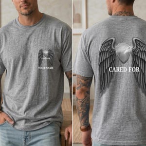 Pode incluir: Camiseta cinza com um desenho de asas de anjo e um escudo nas costas. A frente da camiseta apresenta asas de anjo, o texto "CARED FOR" e "YOUR NAME".