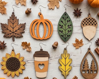 May include: A collection of eight wooden dangle earrings featuring autumn-themed designs including a maple leaf, a pumpkin, a green leaf, an acorn, a sunflower, a coffee cup with a heart, an oak leaf, and a pine tree forest in a teardrop shape.