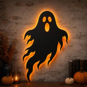 May include: A black ghost-shaped wall decoration with a cutout design for the eyes and mouth, illuminated from behind by a bright orange glow. The silhouette features jagged, flowing edges that mimic a spectral form against a textured wall.