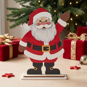 May include: A flat wooden Santa Claus cutout standing on a rectangular base. The figure wears a red suit with white trim, a black belt with a gold buckle, and black boots. Santa has a white beard, a red hat, and one hand raised in a wave.