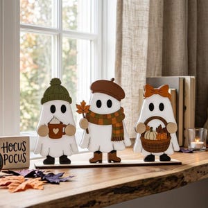 May include: Three white ghost figurines standing on a wooden base. The left ghost wears a green beanie and holds a brown mug with a heart. The middle ghost wears a brown beret and plaid scarf, holding a leaf. The right ghost wears a bow and holds a basket of pumpkins.