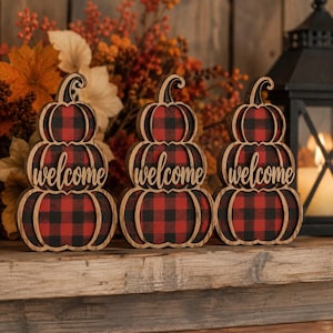 May include: Three stacked pumpkin-shaped wooden signs featuring a red and black buffalo plaid pattern. Each sign has the word welcome written in a cursive script across the centre section. The edges of the pumpkins are finished in a dark wood tone.