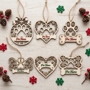 Puede incluir: Seis adornos de madera cortados con láser con formas de huellas, huesos y corazones con detalles de copos de nieve. Cada adorno incluye un cordel de yute y una banda horizontal con el texto impreso Pet Name en tinta roja o verde.