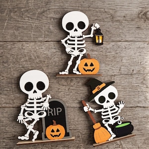 May include: Three wooden Halloween decorations featuring white skeletons with black eye sockets. One skeleton holds a lantern next to a pumpkin, another leans on a tombstone marked RIP with a pumpkin, and the third wears a hat, holding a broom by a cauldron.