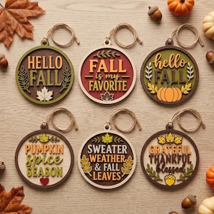 May include: Six circular wooden ornaments featuring autumn-themed designs and text. The ornaments display phrases like HELLO FALL, FALL is my FAVORITE, PUMPKIN SPICE SEASON, SWEATER WEATHER & FALL LEAVES, and GRATEFUL THANKFUL BLESSED, accented with leaves and pumpkins.