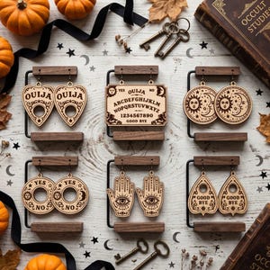 May include: Six pairs of laser-cut wooden earrings displayed on dark wood stands. Designs include heart-shaped planchettes with OUIJA text, a rectangular Ouija board, sun and moon circles, YES NO circles, hamsa hands with eyes, and planchettes with GOOD BYE text.