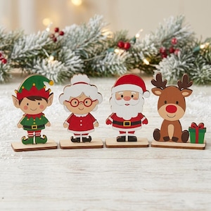 以下が含まれることがあります： 個別の長方形の台座に立った4体の木製クリスマスフィギュアのセット。緑と赤の衣装を着たエルフ、白い髪に赤い眼鏡をかけた女性、赤いスーツを着たサンタクロース、そしてプレゼントを持った茶色のトナカイのフィギュアが含まれています。