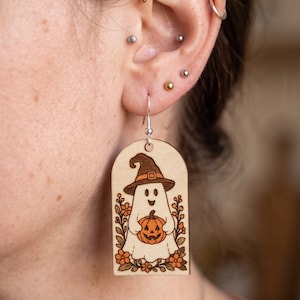 May include: A light-colored wooden dangle earring featuring a laser-engraved illustration of a ghost wearing a witch hat and holding a jack-o'-lantern pumpkin, surrounded by small orange flowers and green leaves, suspended from a silver-toned hook.