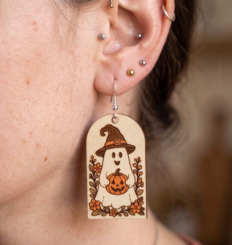 May include: A light-colored wooden dangle earring featuring a laser-engraved illustration of a ghost wearing a witch hat and holding a jack-o'-lantern pumpkin, surrounded by small orange flowers and green leaves, suspended from a silver-toned hook.