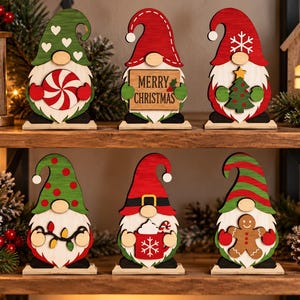 Könnte beinhalten: Sechs flache Holz-Wichtel-Figuren auf rechteckigen Sockeln. Jeder Wichtel hat eine hohe Mütze, einen weißen Bart und ein individuelles Weihnachtsmotiv: ein Pfefferminzbonbon, ein Schild mit MERRY CHRISTMAS, einen Tannenbaum, Lichterketten, einen Becher oder einen Lebkuchenmann.