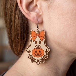 May include: A wooden dangle earring shaped like a ghost wearing an orange bow. The ghost features a carved jack-o'-lantern pumpkin in its center, surrounded by spider web details and a small spider at the bottom, hanging from a dark metal hook.
