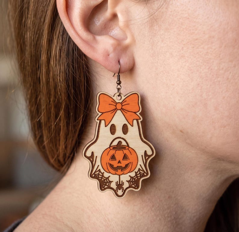 May include: A wooden dangle earring shaped like a ghost wearing an orange bow. The ghost features a carved jack-o'-lantern pumpkin in its center, surrounded by spider web details and a small spider at the bottom, hanging from a dark metal hook.