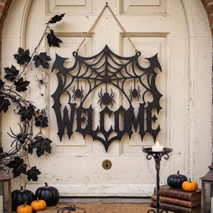 Puede incluir: Un letrero colgante de madera negra con un diseño de telaraña con tres arañas y la palabra WELCOME en una fuente gótica. El letrero está suspendido por una cuerda de yute y decorado con hojas negras, calabazas negras y una vela en un soporte.