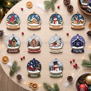 May include: A collection of ten wooden snow globe ornaments featuring various holiday scenes including a cardinal, snowman, gingerbread house, red truck, and nativity. Each ornament displays text such as Season's Greetings, Let it Snow, and O Holy Night.