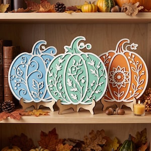 May include: Three decorative wooden pumpkin cutouts displayed on small stands. The left pumpkin is light blue, the center is mint green, and the right is orange. Each features intricate white laser-cut floral and vine overlays on the front.