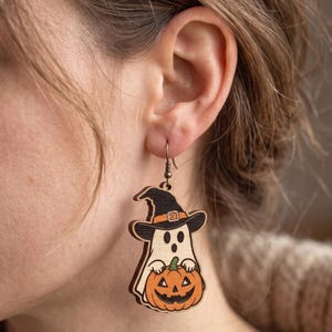 May include: A wooden dangle earring featuring a light-colored ghost wearing a black witch hat with an orange band. The ghost is holding an orange jack-o'-lantern with a black carved face. The earring hangs from a simple metal hook.